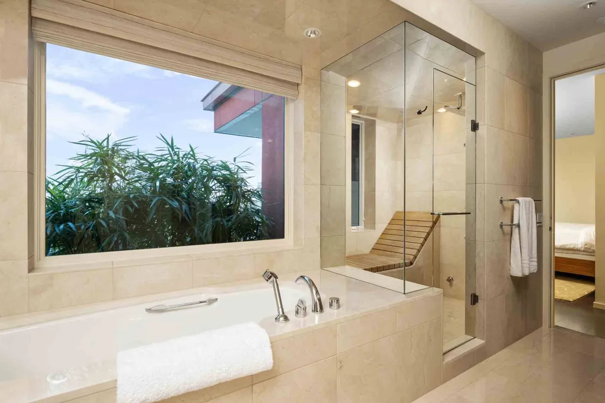 A modern bathroom featuring a luxurious bathtub and a shower enclosure next to a large window with lush greenery outside. The space is calm and rejuvenating, with daylight illuminating the serene setting.