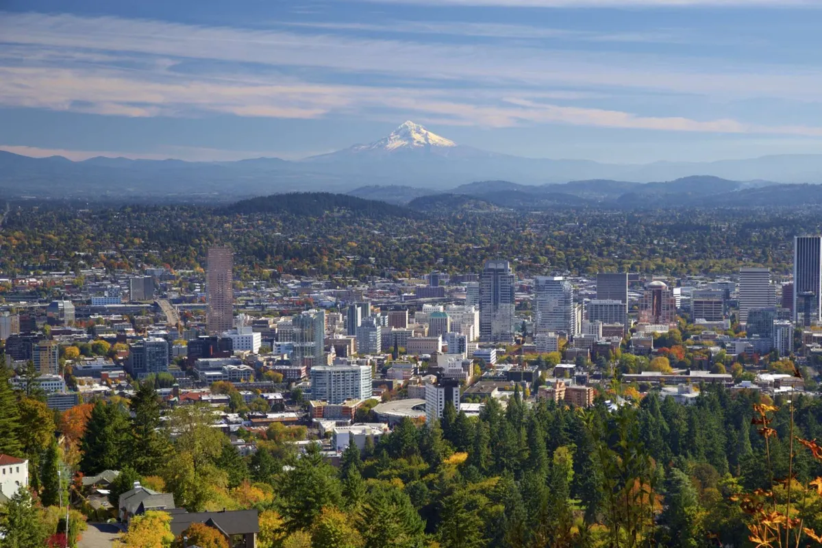 A sweeping, high-angle view over Portland featuring the city skyline, lush greenery in the foreground, and the iconic snow-capped Mount Hood in the distance. The scene merges urban excitement with natural beauty, conveying the city's unique blend of metropolitan life and access to green spaces.