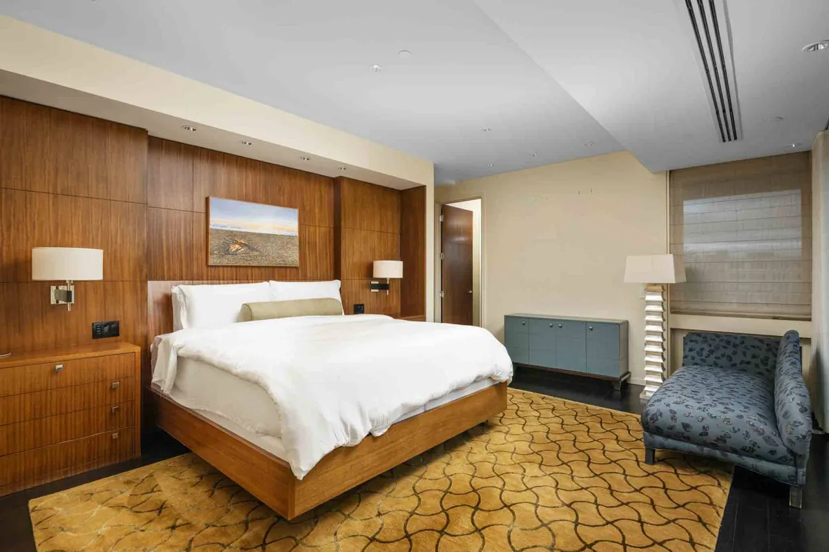 A tranquil bedroom with a comfortable bed, soft natural light streaming through the windows, a plush sofa at the foot of the bed, contemporary artwork, and cozy textures throughout. The overall feel is peaceful and welcoming.