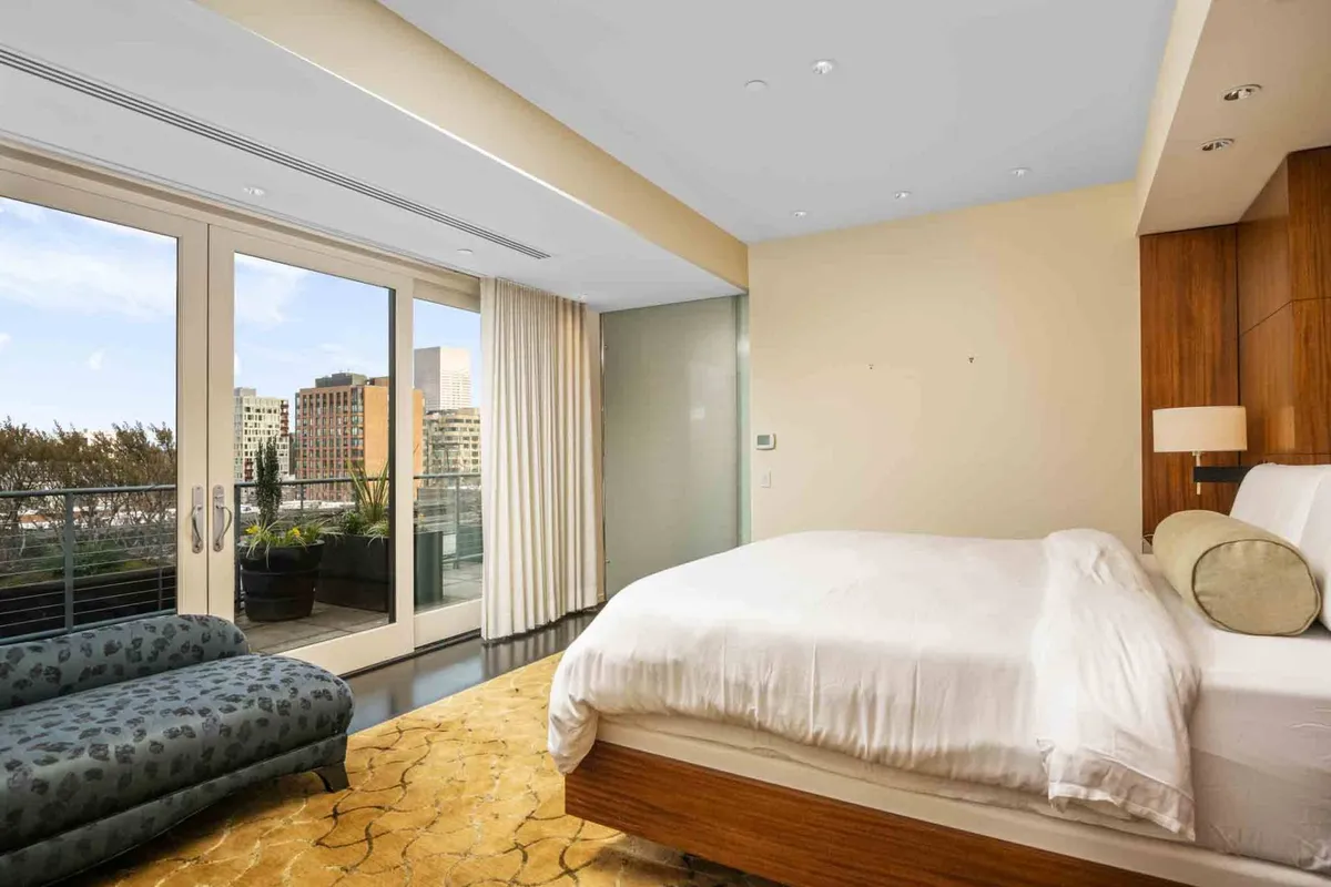 A serene and luxurious bedroom with a large bed dressed in white linens, a stylish chaise lounge, modern furniture, and floor-to-ceiling windows that frame sweeping city views. Natural light pours in, creating an inviting and restful atmosphere.