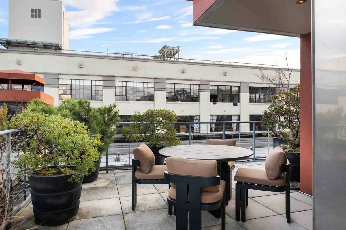 A tranquil, well-appointed outdoor seating area framed by lush green plants, with an urban backdrop stretching into the distance. The setting captures both relaxation and social possibilities, inviting morning coffee or evening cocktails with a view.