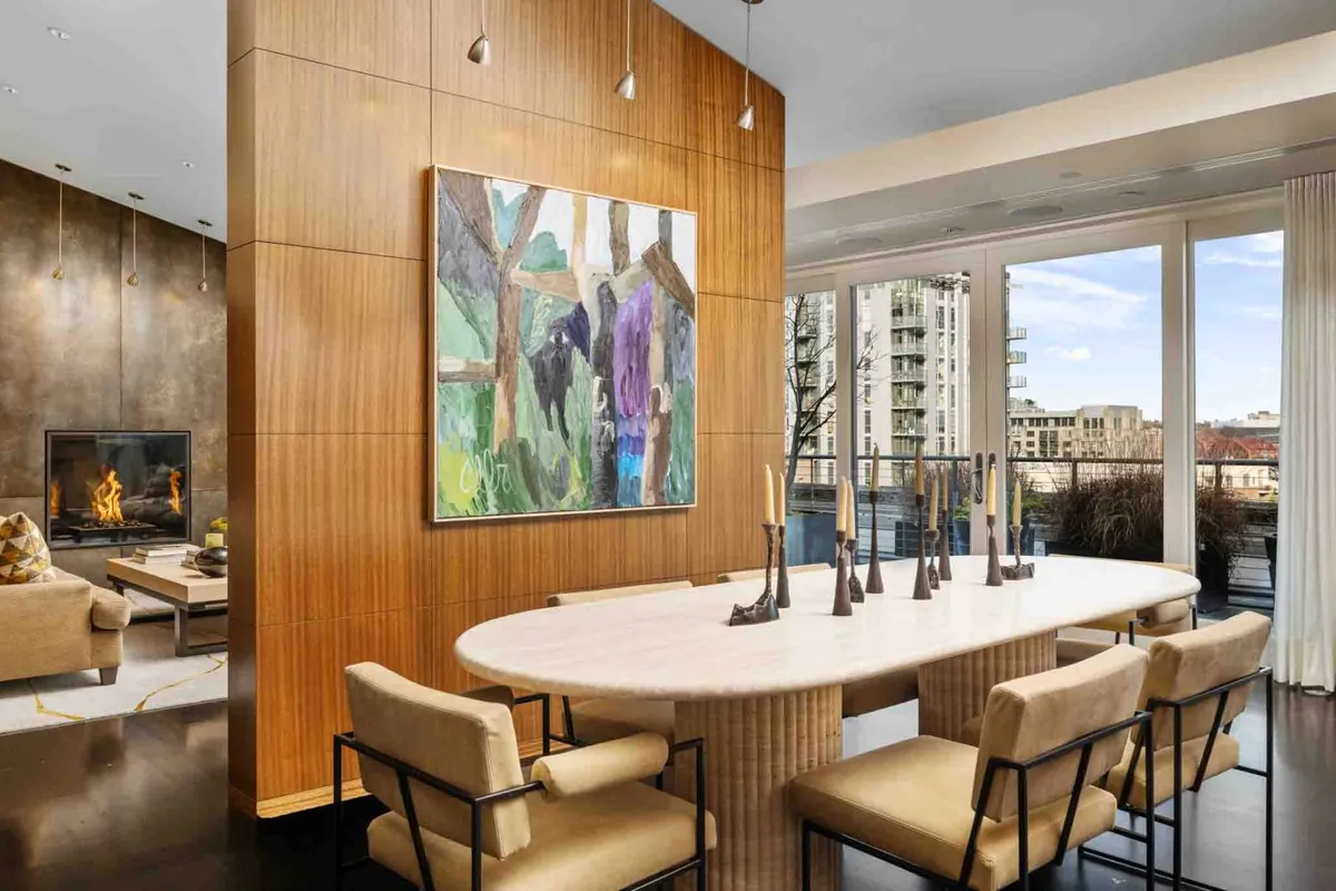 This image captures an elegant dining area featuring a large dining table, striking colorful artwork, a modern fireplace, and expansive floor-to-ceiling windows that offer a dramatic view of the city skyline.