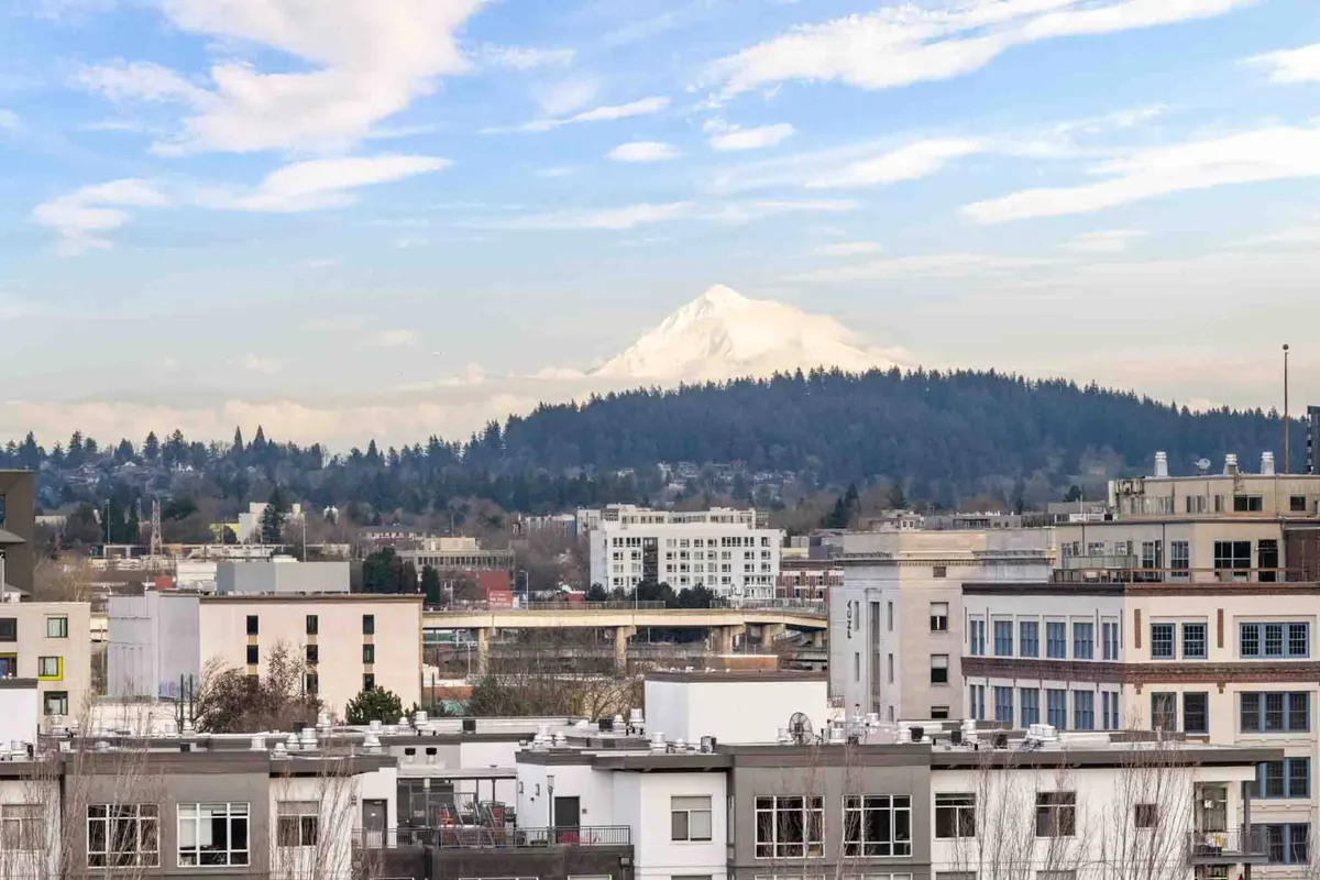 The view showcases Mount Hood rising beyond Portland’s urban skyline beneath layered clouds. The composition highlights both the natural beauty and the cosmopolitan feel of the city, suggesting close proximity to both outdoor adventures and vibrant city life.