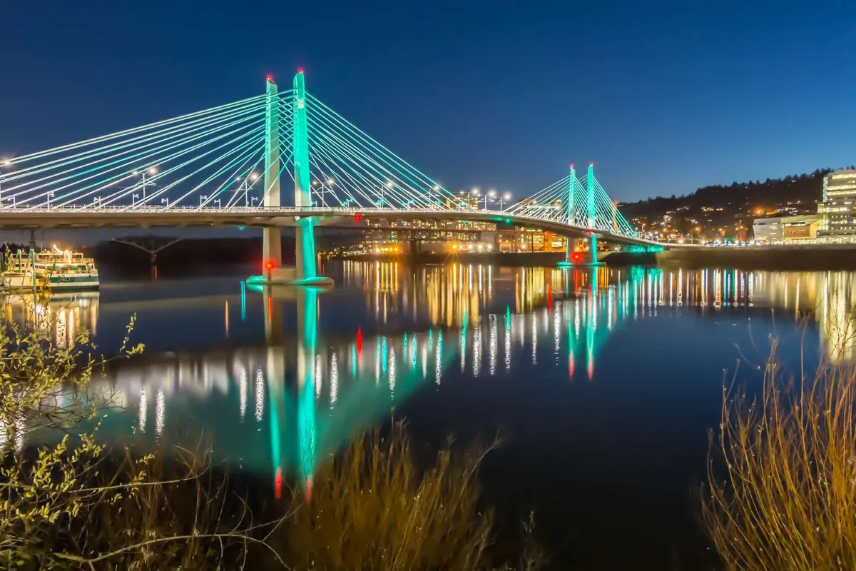 A nighttime exterior scene featuring an illuminated bridge with city lights reflecting across the water, signaling the vibrant nightlife and riverfront activity nearby.