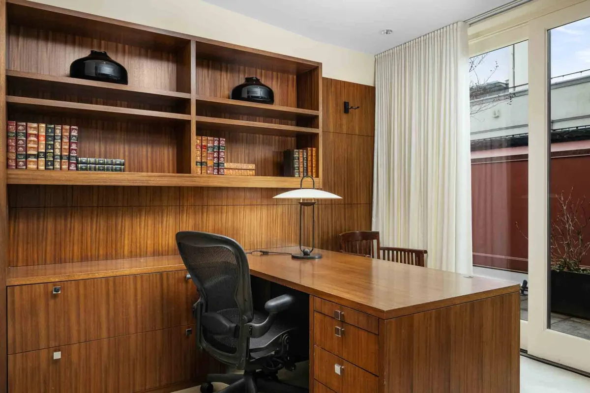 A serene office space featuring a wooden desk set against a backdrop of large windows that flood the room with natural light. A bookshelf neatly arranged with books and a classic desk lamp create an atmosphere tailored for focused work, study, or creative thought.