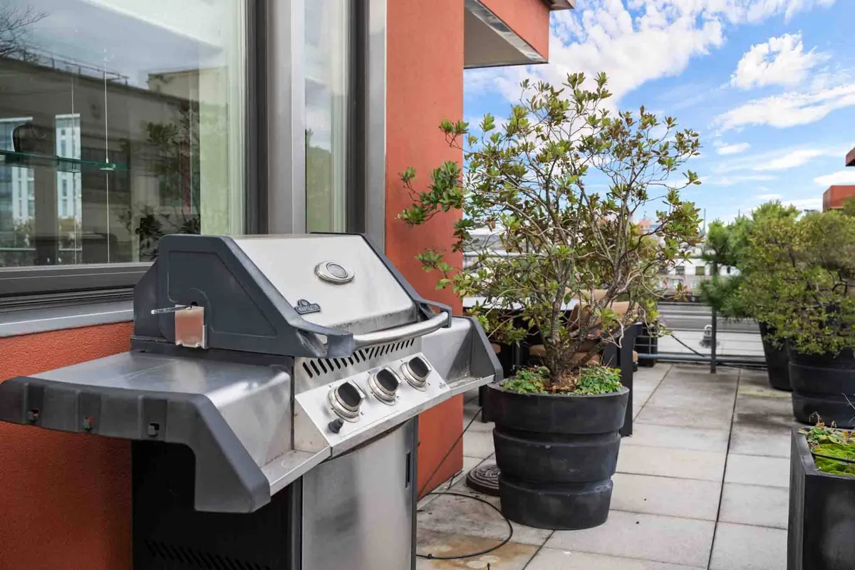 A private balcony set up for outdoor dining with a grill and potted plants, looking out over the city. The scene is relaxed and social, suggesting morning coffee or sunset barbecues with friends.