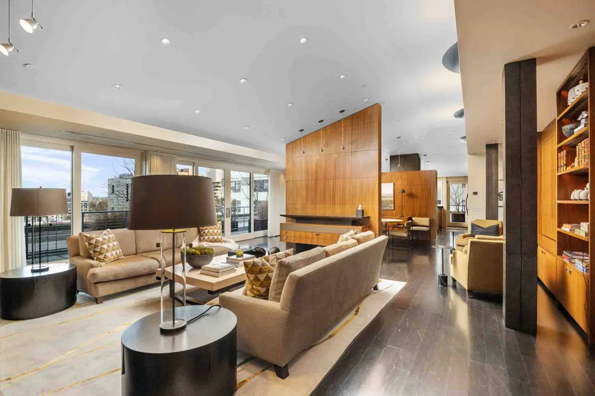 The image showcases a spacious, light-filled penthouse living room featuring contemporary sofas, a stylish coffee table adorned with decorative pieces, a striking wooden feature wall, and expansive windows that flood the space with natural light and offer city views. The setting feels inviting and luxurious, perfect for relaxing, socializing, or enjoying quiet moments with a book.